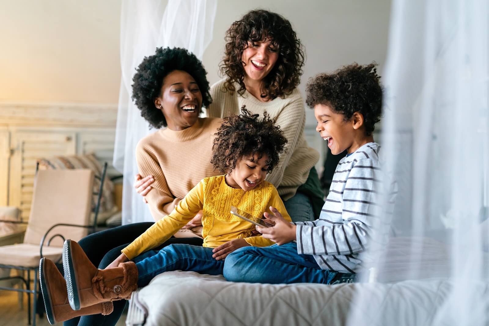 A biracial LGBTQ+ couple sitting & laughing with their children on a bed. Representing how IHS can support the whole family. Learn how LGBTQIA+ therapy in Pelham, AL can help your family thrive.