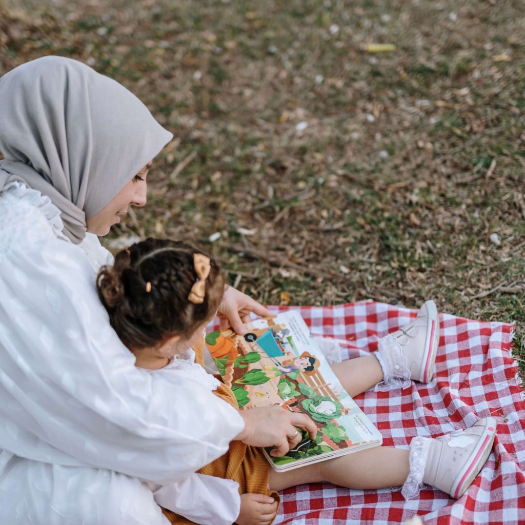 A mother sitting with her child, reading a book & sitting on a red gingham blanket. Postpartum therapy in Alabama is here to support mothers navigating postpartum challenges. Learn more here about working with a therapist. 