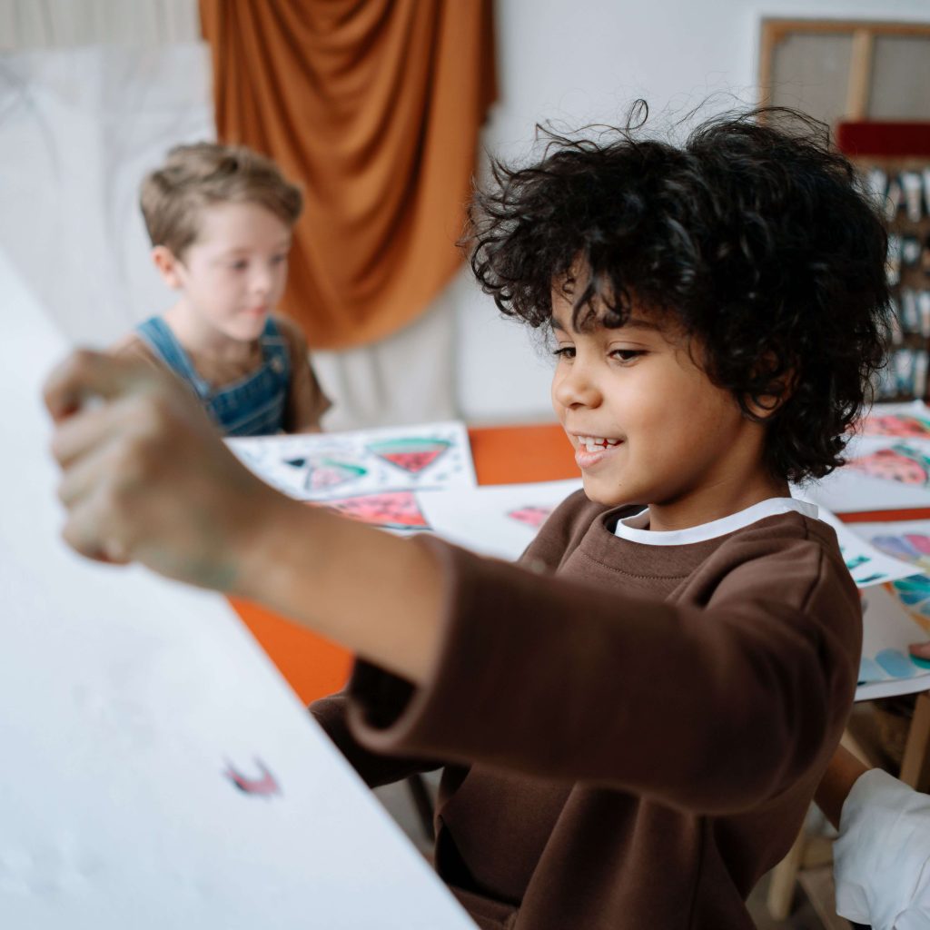A child smiling while holding up their artwork near a friend. Discover how neurodivergent affirming therapy in Pelham, AL can support yourself, your child or teenager. Learn more here.
