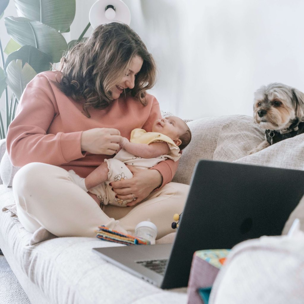 A happy mom sitting with a newborn baby while sitting near a laptop & small dog. Representing working moms & how postpartum therapy in Birmingham, AL can support your mental health. Find a postpartum therapist today.